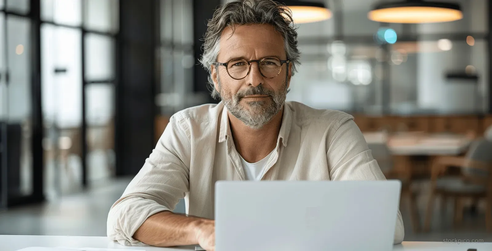 A middle-aged man with glasses types on a laptop at an office desk with blurred windows behind him.
