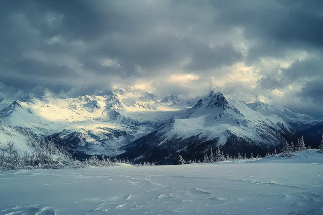 Snow-covered mountains stretch beneath dark clouds while pale light breaks through over a winter forest valley.