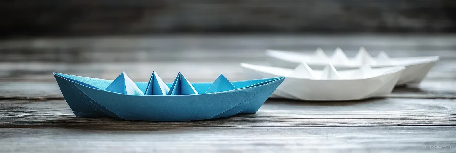 A blue origami boat leads several white paper boats on a vintage wooden table.