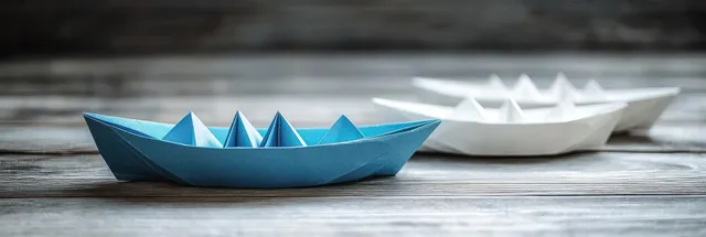 A blue origami boat leads several white paper boats on a vintage wooden table.