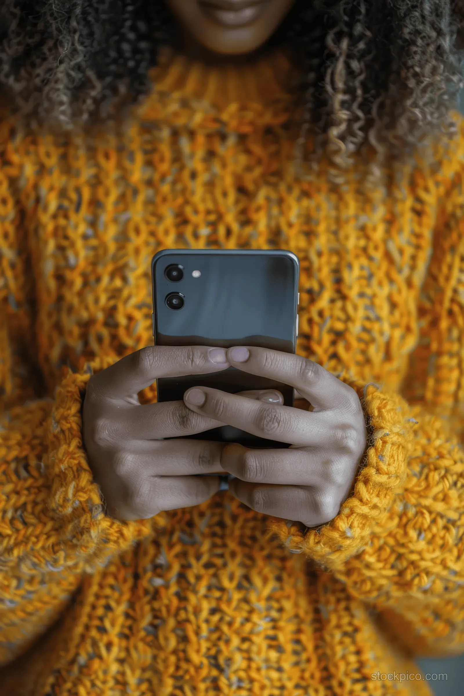 A woman in a yellow knit sweater holds a smartphone and types a message indoors.