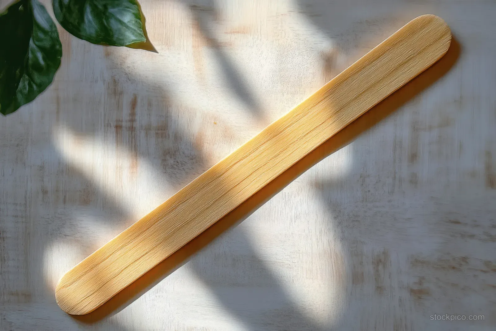 A wooden tongue depressor rests on a light wood table with soft sunlight shadow and green leaves in the corner.