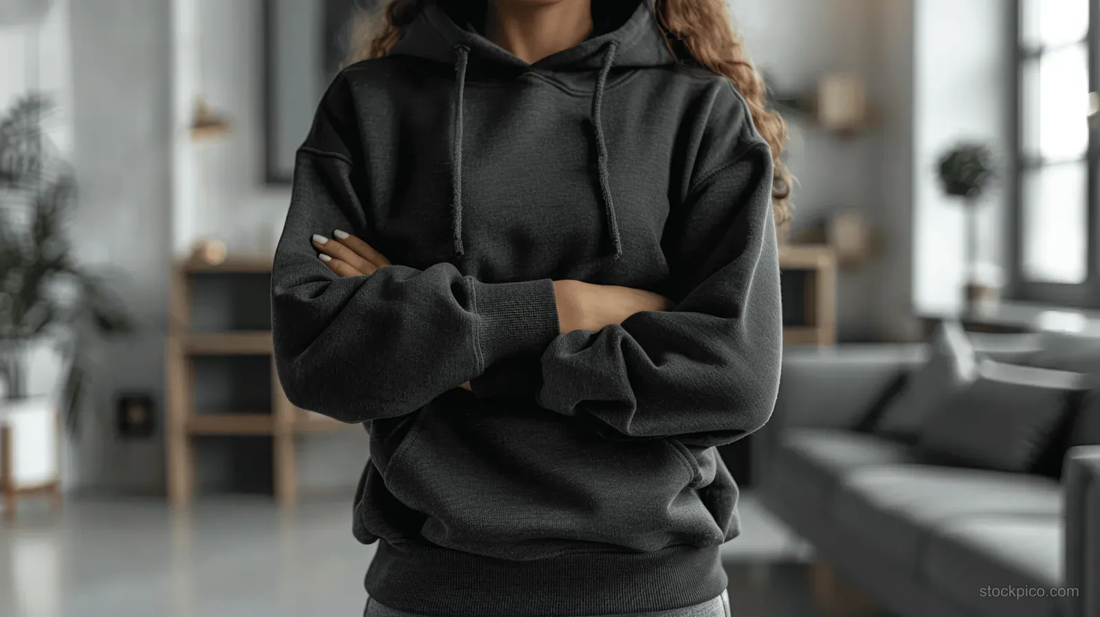 Person in a black hoodie with arms crossed, standing indoors in a bright living room near windows.