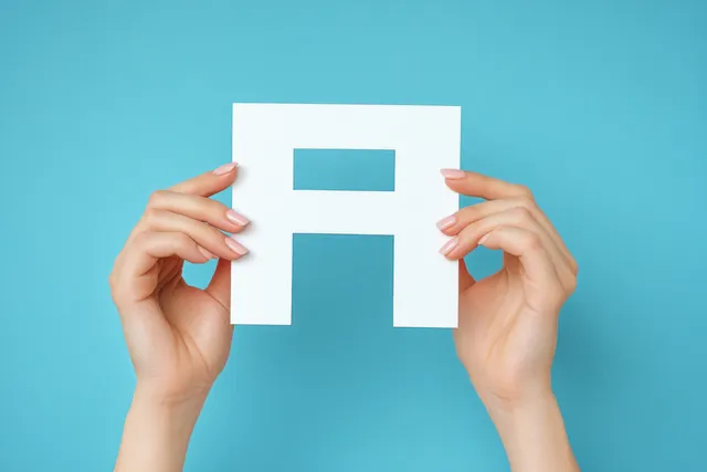 Two hands hold a white paper cutout capital letter centered against a solid blue background.