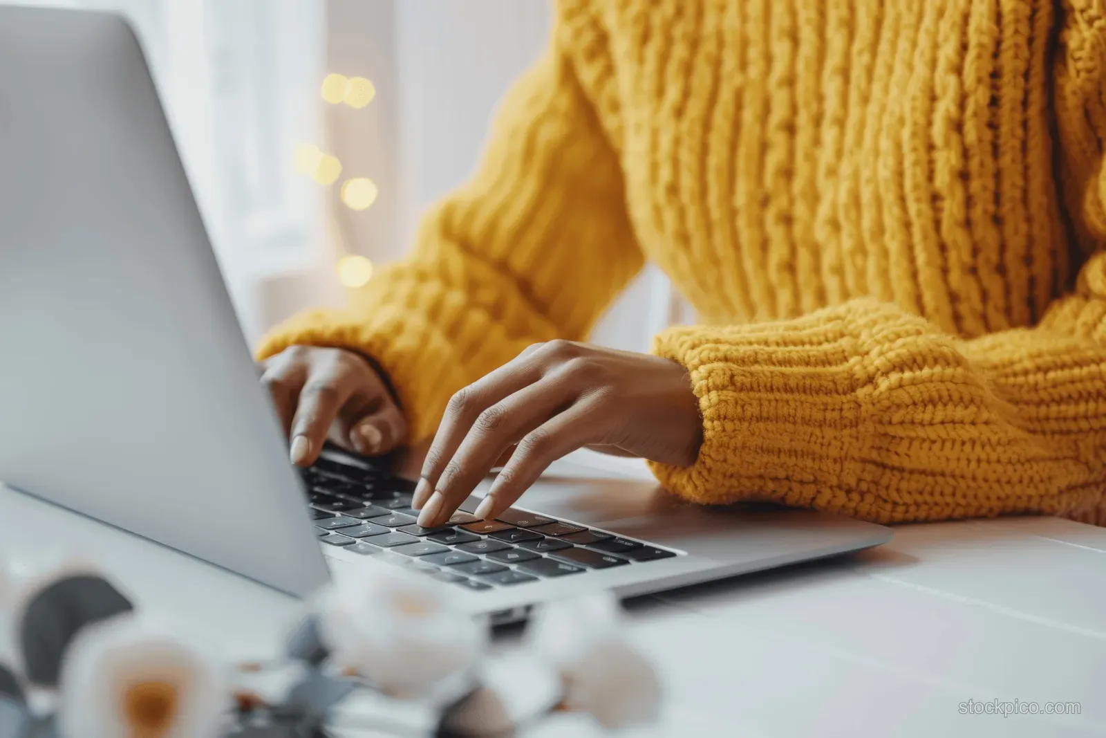 Woman’s hands typing on a laptop in a bright minimalist white office, with blurred desk items in the foreground.
