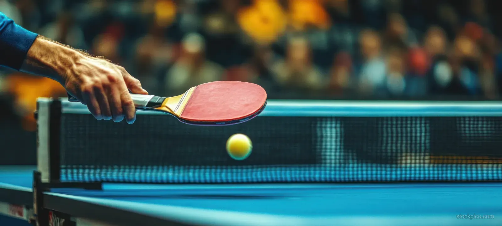 Close-up of a red paddle hitting a yellow ping pong ball over a blue table with the net and blurred crowd behind.