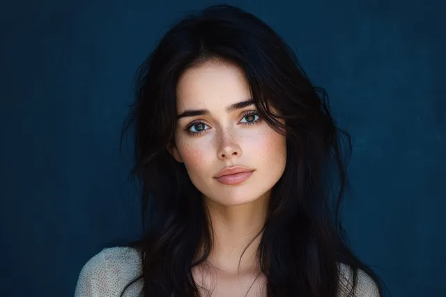 Young woman with freckles and long dark hair gazes softly into camera against dark blue