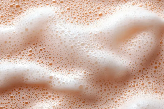Pastel pink and beige foam bubbles spread over a skincare cleanser surface in close-up.