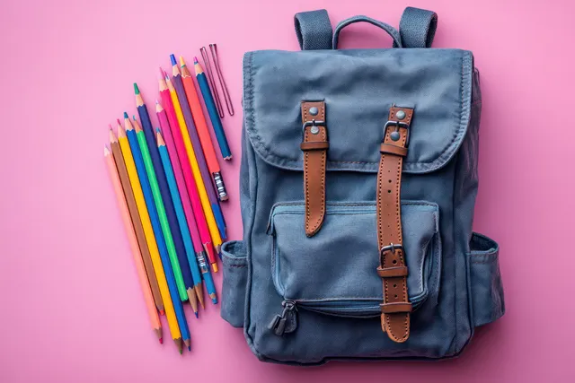 Gray backpack on a pink background with colorful pencils and notebook stationery arranged in a flat lay.