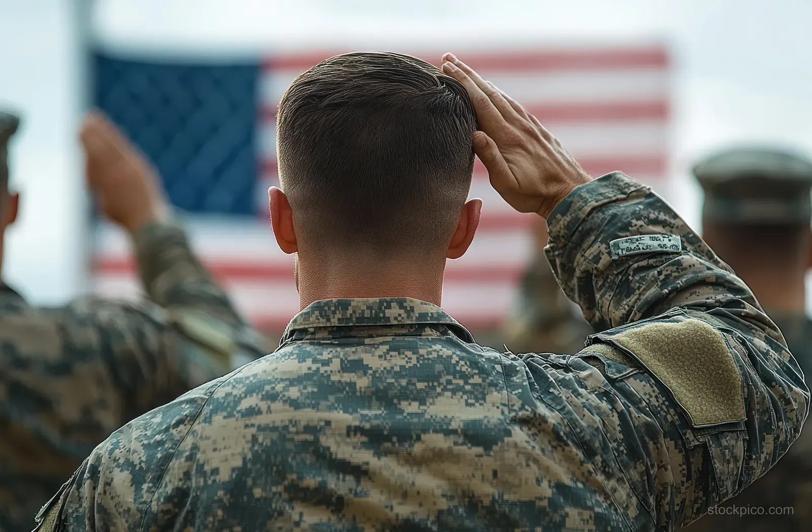 Soldier in camouflage salutes with his back to camera in front of an American flag.