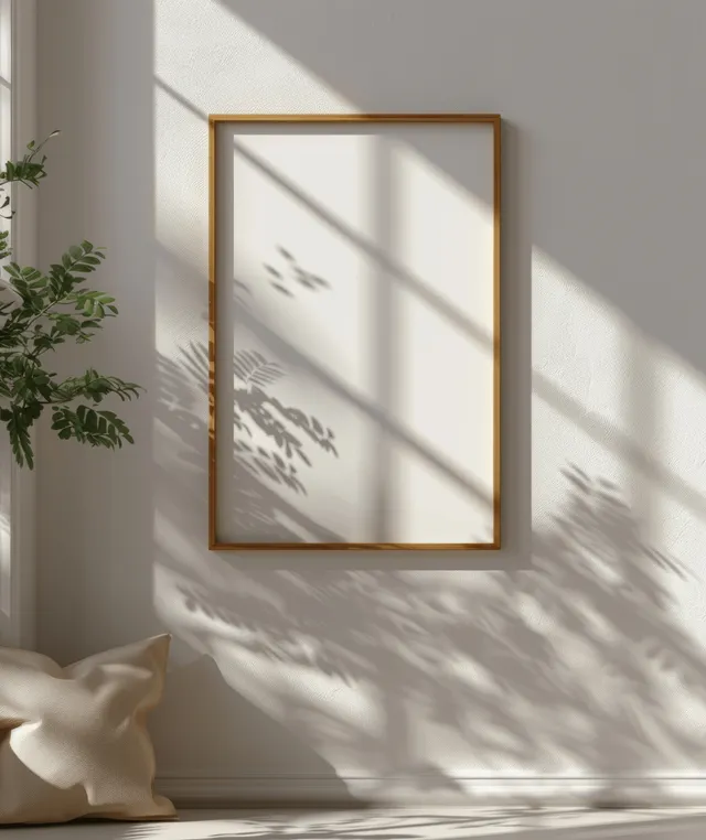 A vertical wooden picture frame hangs on a neutral wall with soft window-shadow stripes across it.