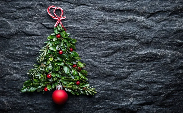 Mini Christmas tree with red ornament and ribbon bow placed on dark textured stone, with candy canes beside it.