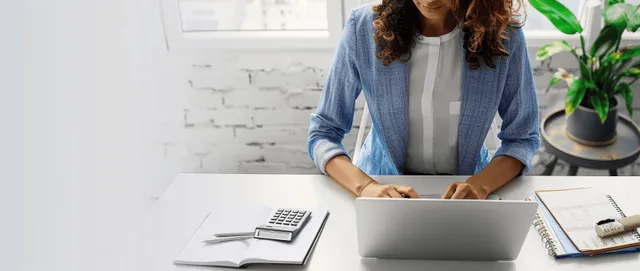 Businesswoman at a desk using a laptop and calculator next to a notebook and financial papers in a bright office.