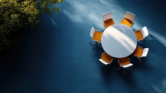 Top-down view shows a round white table encircled by eight orange and white chairs on a blue floor with sunlight shadows.