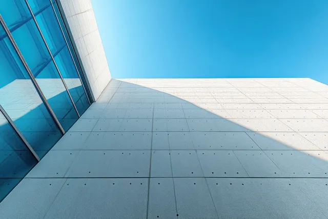 White concrete building facade with rectangular glass windows reflecting the blue sky and casting sharp shadows.
