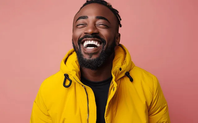 Laughing young Black man in yellow hoodie smiling against pastel pink backdrop