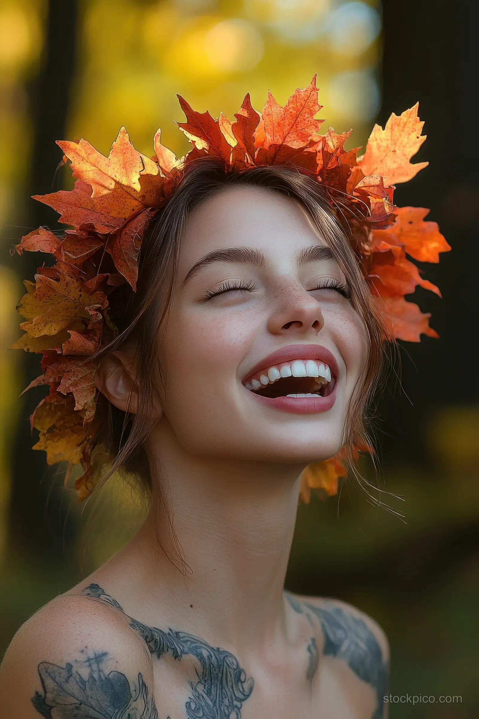 A joyful young woman smiles with eyes closed wearing a vibrant autumn leaf crown outdoors in warm sunlight.