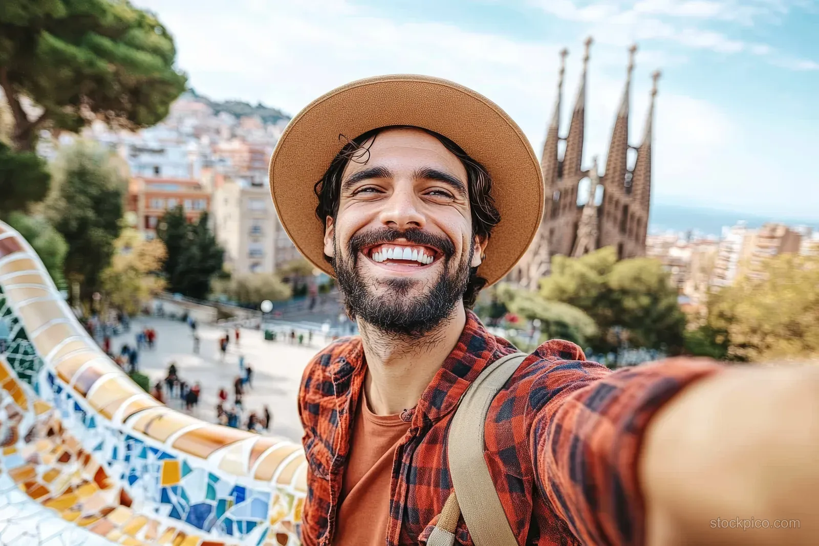 Man taking a selfie on Park Güell mosaic terrace with Sagrada Família visible over Barcelona buildings.