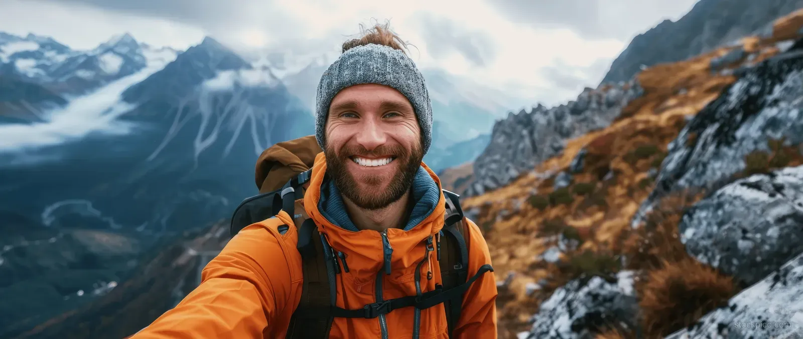 Smiling hiker with backpack taking a selfie on a snowy mountain summit with rocky slopes and distant peaks.