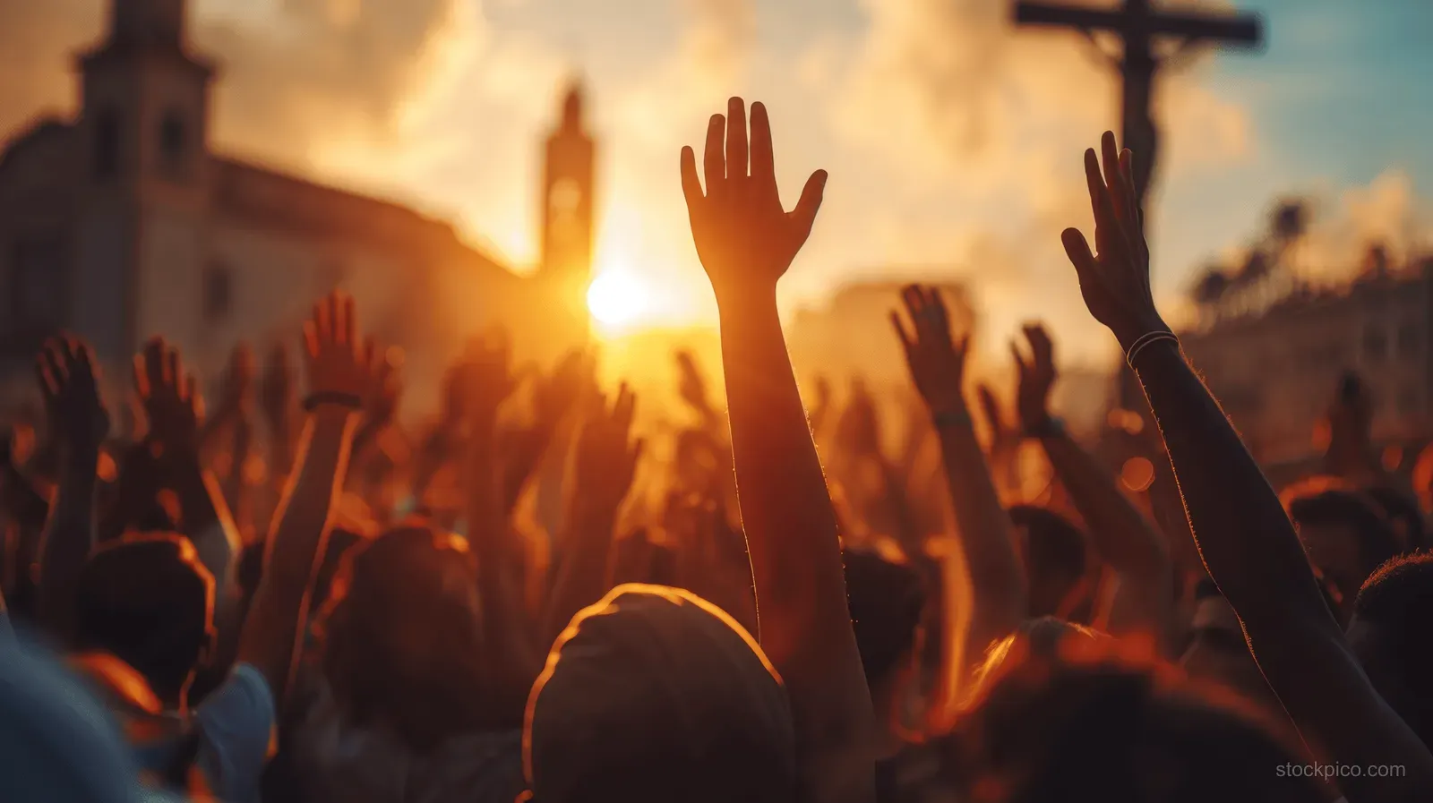 A crowd raises hands in prayer at sunset while a cross silhouette stands behind them.