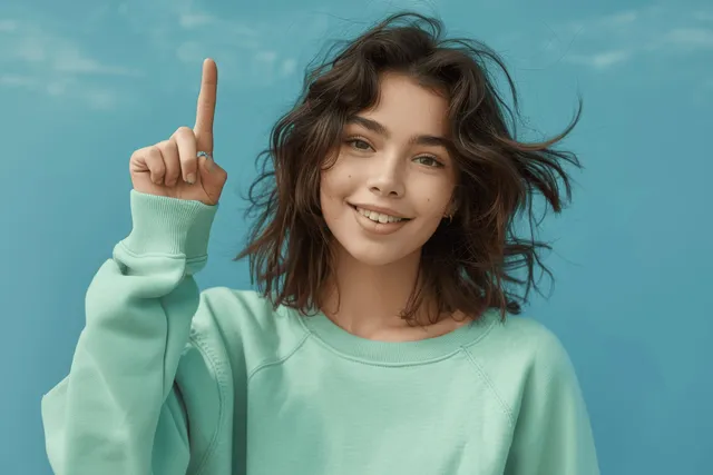 Young woman smiles while raising index finger in light green sweatshirt against blue background