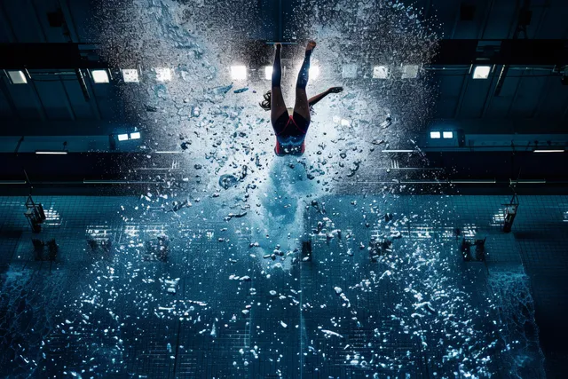 A female diver leaps from a 3m springboard in an indoor pool, with splash clouds bursting below her.