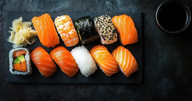 Assorted sushi nigiri and rolled pieces on a black slate plate with soy sauce and pickled ginger.