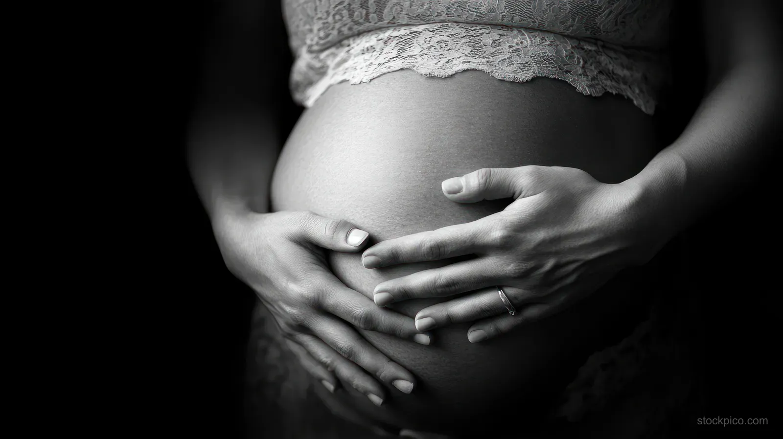 Monochrome close-up of a pregnant mother’s hands cradling her belly under soft diffused light.