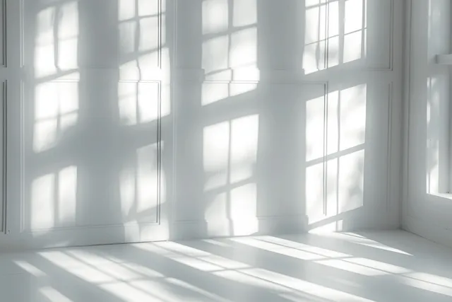 Sunlight through a window creates soft layered shadow overlays on a white minimalist room floor.