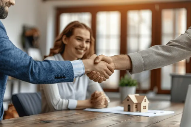A realtor shakes hands with a couple at a desk with contract papers and a small house model.