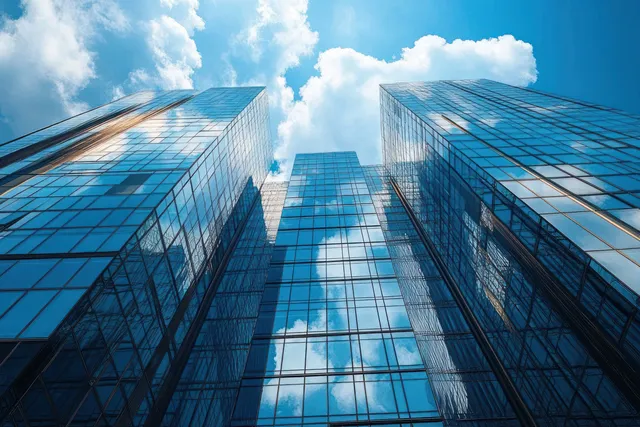 Low-angle view of tall glass skyscrapers reflecting fluffy clouds under a bright blue sky.