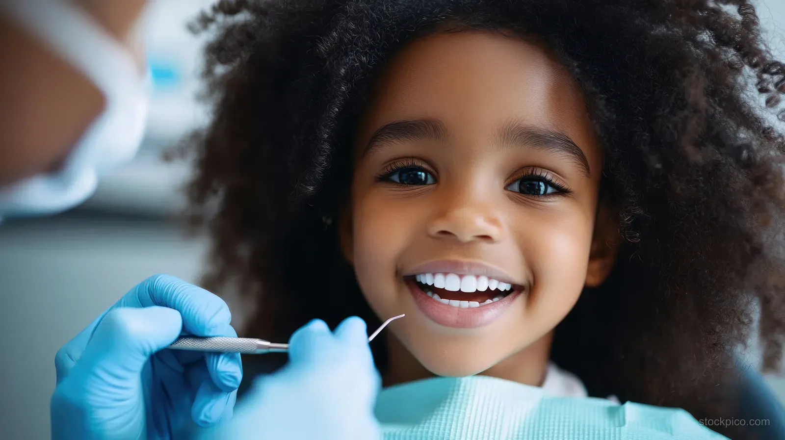 A smiling girl with curly hair has her teeth examined by a dentist wearing blue gloves in a dental clinic.