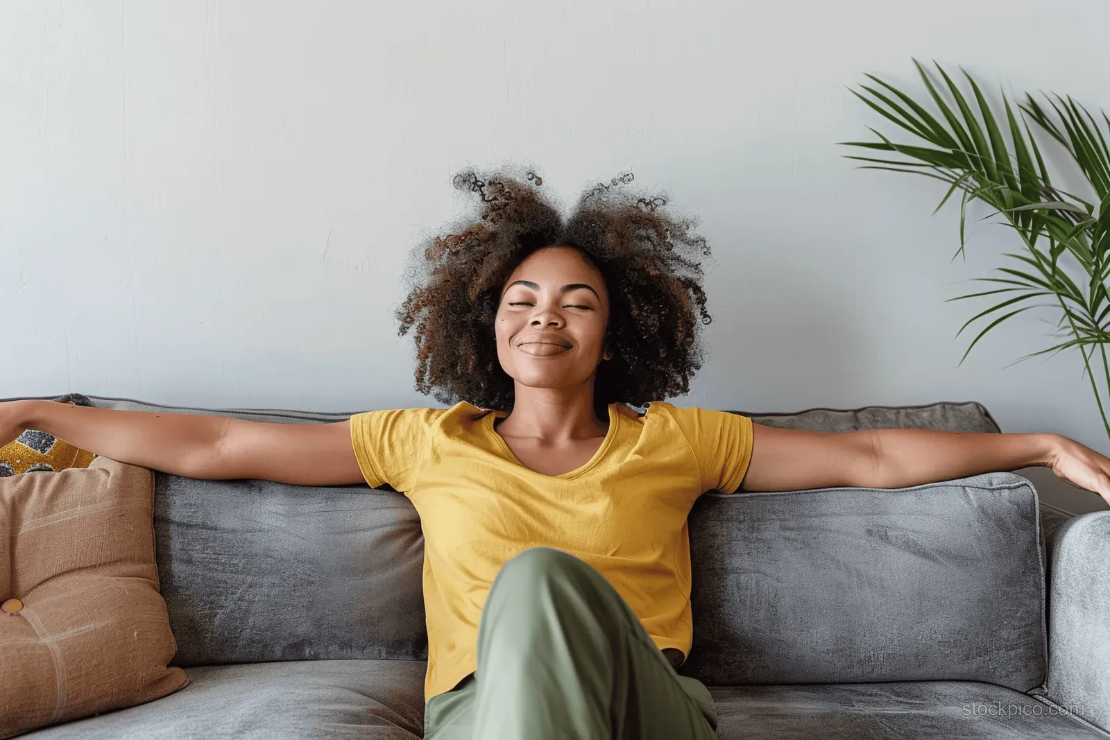 Smiling woman stretches on a gray sofa in a minimalist living room with white wall and a plant.