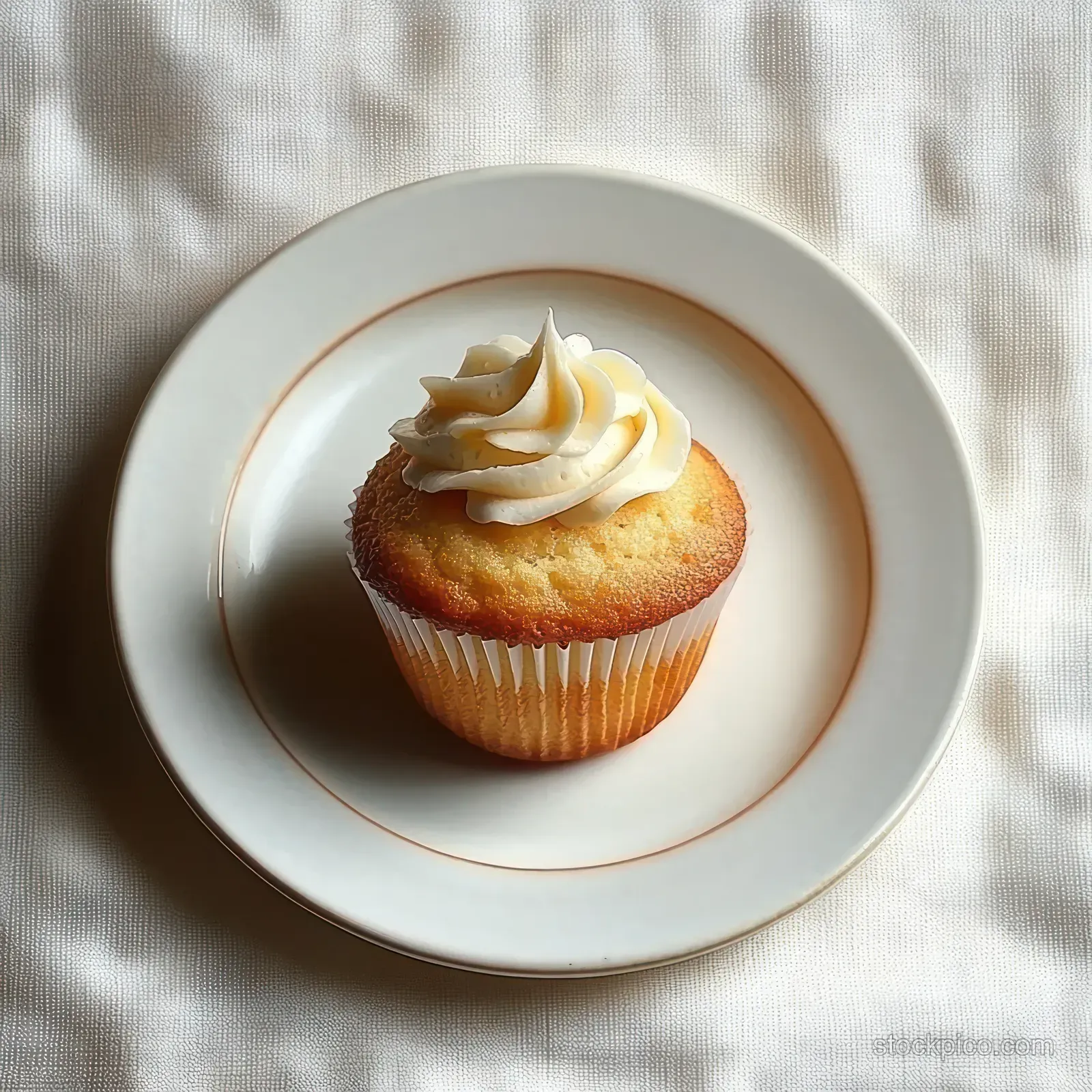 A single vanilla cupcake with white frosting rests on a white plate with a gold rim on textured white fabric.