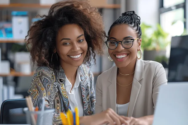Two businesswomen smile while discussing laptop plans at a desk in a bright office.
