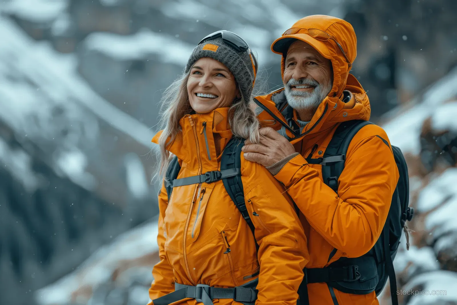 Elderly man smiling at the camera while an older woman hikes beside him through snowy mountains.