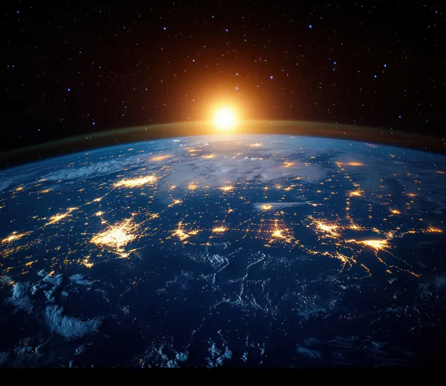 Earth seen from space at night with city lights and a sunrise rim lighting the atmosphere behind stars.