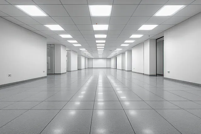 Empty office corridor with white walls, bright ceiling light panels, gray tiled floor, and open doorways.