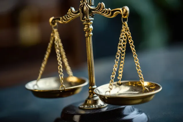 Golden balance scale with chains on a stone base, softly lit with reflections in a courtroom setting.