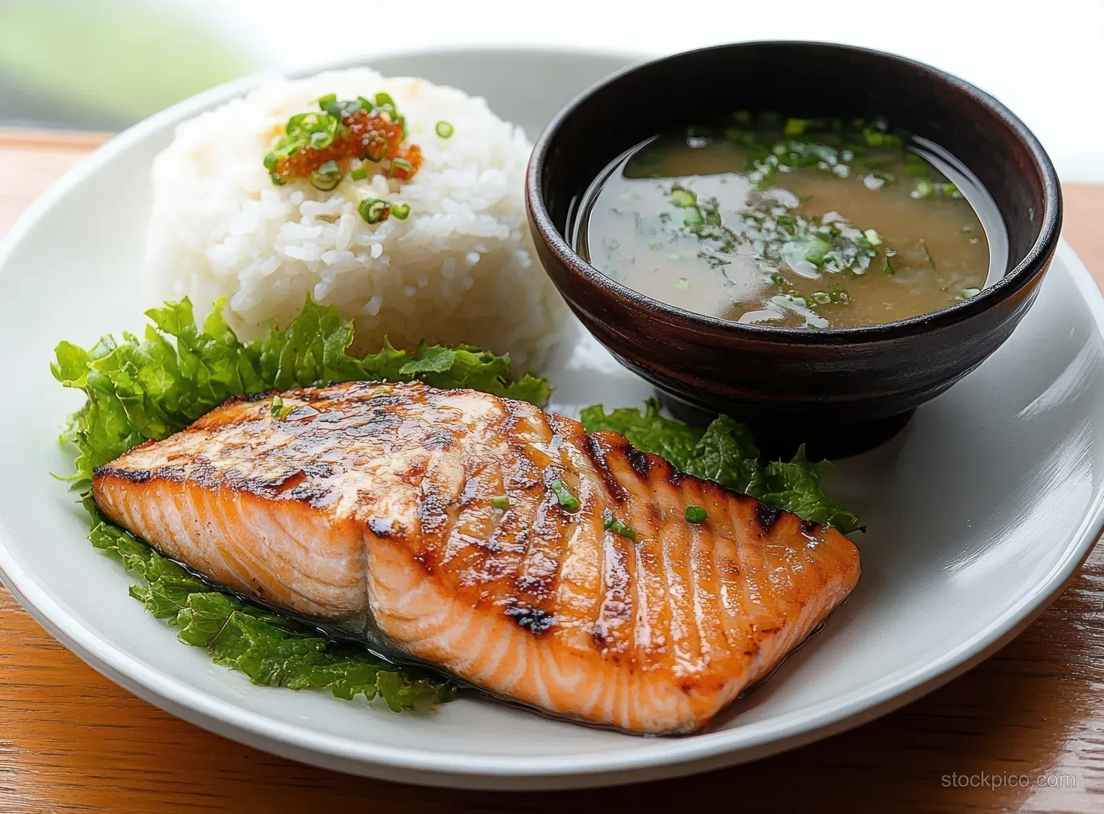 Grilled salmon fillet on lettuce next to white rice with green onion and a bowl of clear herb soup on a white plate.