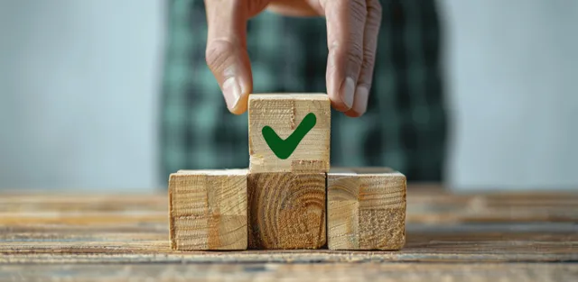 A hand places a green checkmarked block atop wooden blocks to show compliance success.