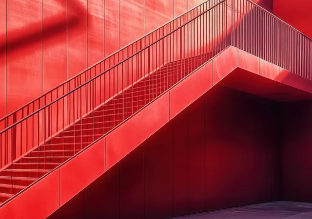 A red staircase with metal railing casts geometric shadows on a paneled red wall in bright daylight.