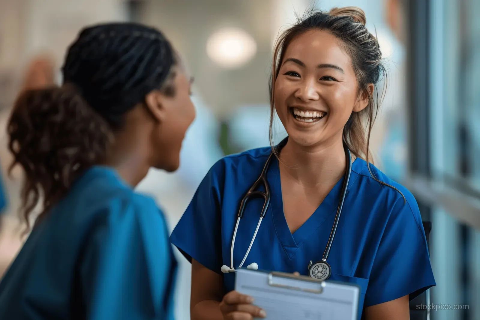 Smiling nurse in blue scrubs talks with another nurse in a hospital hallway while holding a clipboard.