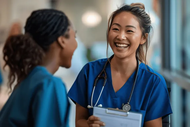 Smiling nurse in blue scrubs talks with another nurse in a hospital hallway while holding a clipboard.