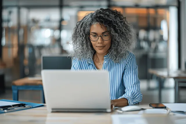 A woman in glasses types on a laptop at a desk in a glass-walled modern office.