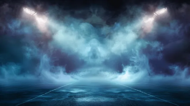 Blue stadium spotlights beam through thick fog above an empty sports field at night.