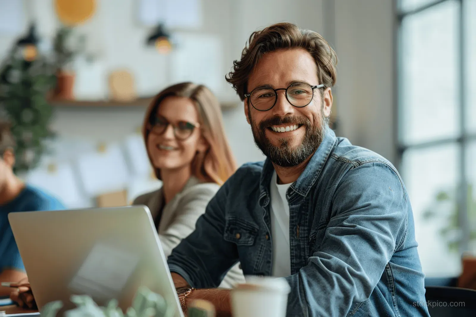 Smiling businessman and female colleague discuss business plan in a bright office while seated with laptops.