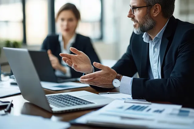 An executive gestures beside laptop screens in a boardroom while colleagues listen behind him.
