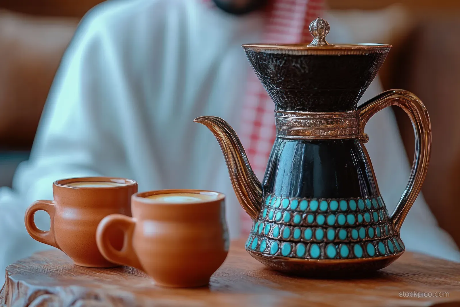 Ornate traditional coffee pot with turquoise detailing beside two brown ceramic cups on a wooden table.