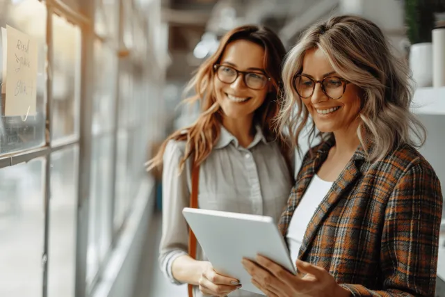 Two women brainstorming business ideas in an office hallway, reviewing a tablet together